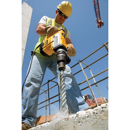 A construction worker wearing a yellow safety helmet and gloves is using a DEWALT rotary hammer drill (model DT9410) to drill into concrete at a construction site. The worker is standing on a concrete surface, and rebar and a crane are visible in the background against a clear blue sky.