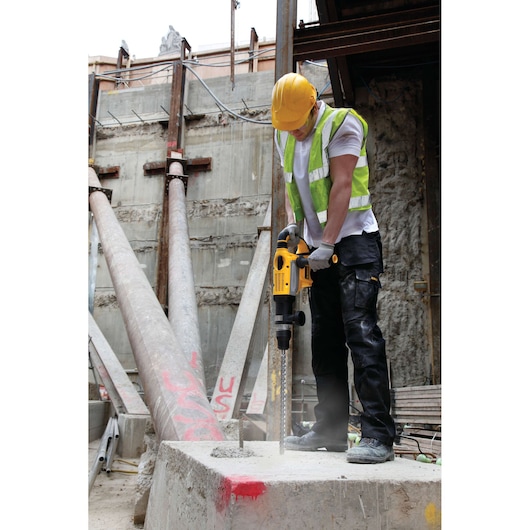 A construction worker wearing a yellow hard hat, high-visibility vest, gloves, and work pants operates a yellow DEWALT rotary hammer drill on a concrete block at an outdoor construction site.