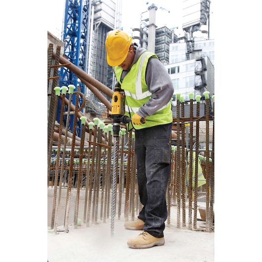 A construction worker wearing a yellow hard hat and reflective vest is using a DEWALT power tool to drill into concrete at a building site. Steel rebar with green caps surrounds the worker, and modern buildings and cranes are visible in the background.