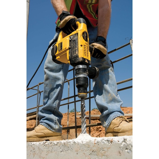 A person using a yellow DEWALT rotary hammer drill to bore into a concrete surface at a construction site. The drill is powered by a cord, and the person is wearing jeans, work boots, gloves, and a reflective vest.