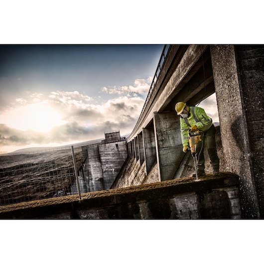 A construction worker wearing safety gear uses a DEWALT power tool to drill into concrete on a large industrial structure