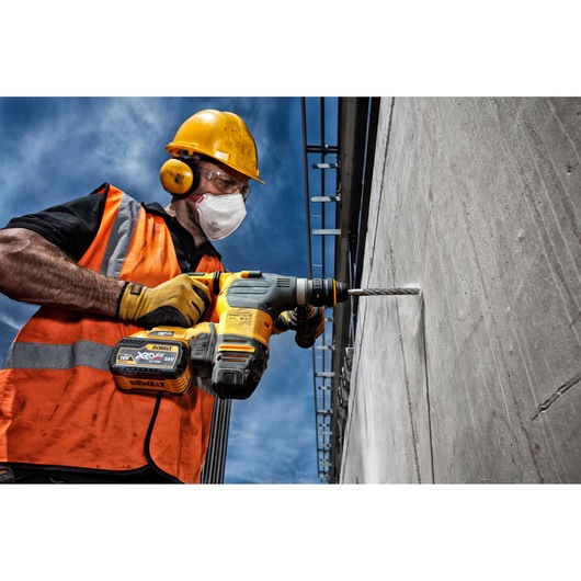 A construction worker wearing an orange safety vest, yellow hard hat, and gloves is using a DEWALT cordless rotary hammer drill with an XR FLEXVOLT battery to drill into a concrete wall, outdoors under a blue sky.
