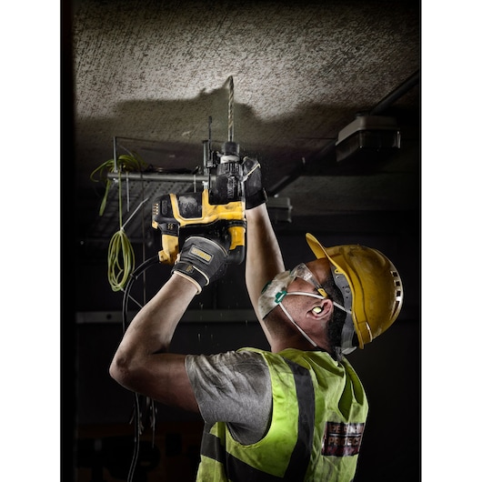 A construction worker wearing a yellow hard hat, safety vest, gloves, ear protection, and face mask is using a DEWALT power drill to drill into a ceiling. Power cables and building structure are visible in the background.