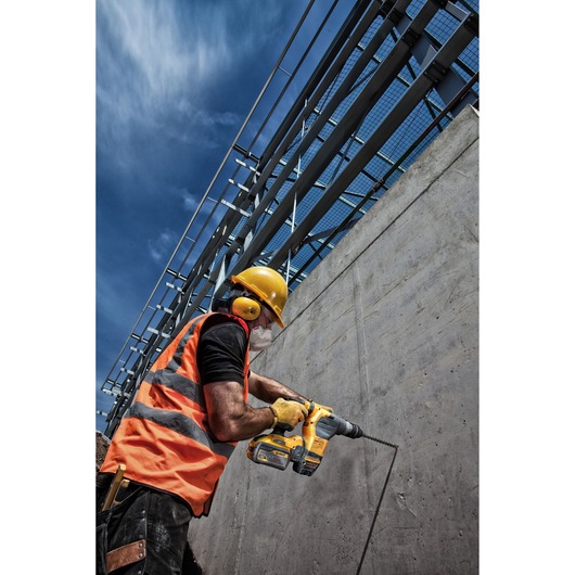 A construction worker wearing a yellow hard hat, orange safety vest, yellow ear protection, and gloves is using a DEWALT cordless hammer drill to drill into a concrete wall at a construction site under a blue sky.