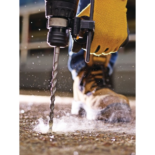 Close-up of a person wearing a yellow work glove using a power drill with a masonry bit to drill into a concrete surface, with dust and debris scattering.