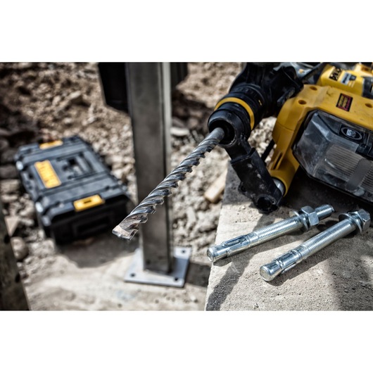 Close-up of a DEWALT yellow power drill with a large masonry drill bit attached, placed on a concrete surface next to three metal anchor bolts. In the background, there is a DEWALT carrying case and construction materials.
