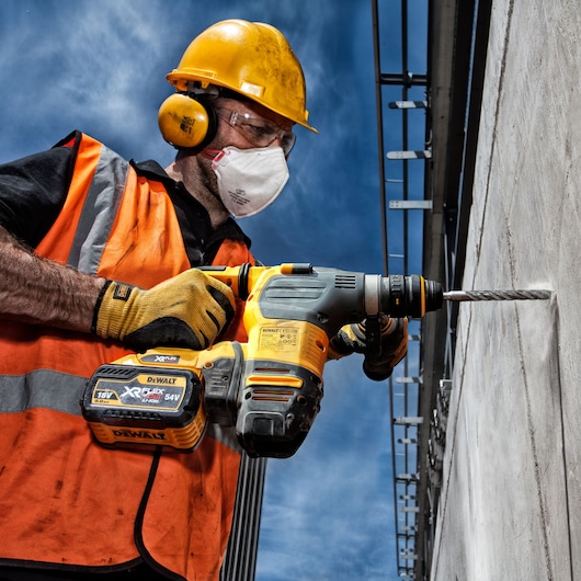 A construction worker wearing a yellow hard hat, orange safety vest, and protective gloves is using a DEWALT DT9580 cordless power drill to drill into a concrete wall. The scene is outdoors with a blue sky in the background.