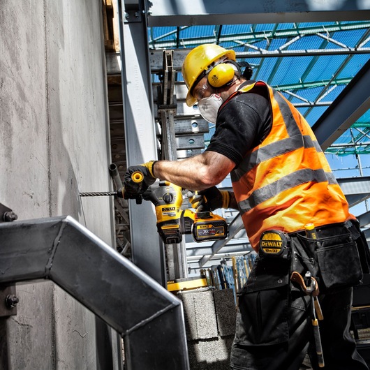 Construction worker wearing protective equipment and an orange safety vest drills into a concrete wall with a DEWALT cordless rotary hammer drill. The worker is on a building site with steel beams and blocks visible around.