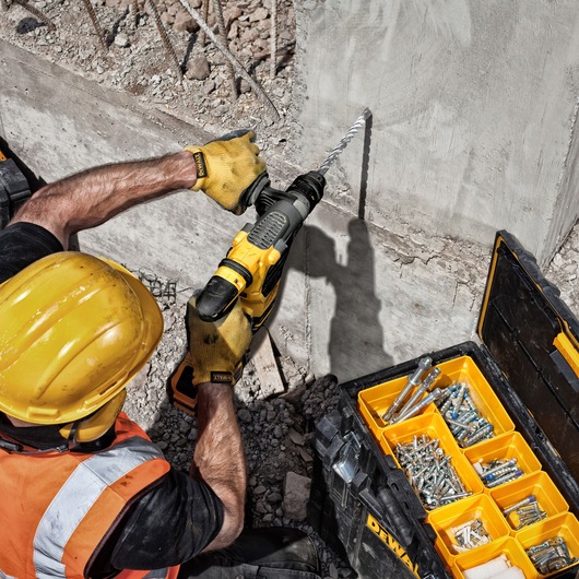 A construction worker wearing a yellow hard hat and gloves uses a DEWALT drill to make a hole in a concrete wall. Next to the worker is an open DEWALT tool box containing various screws and fasteners.