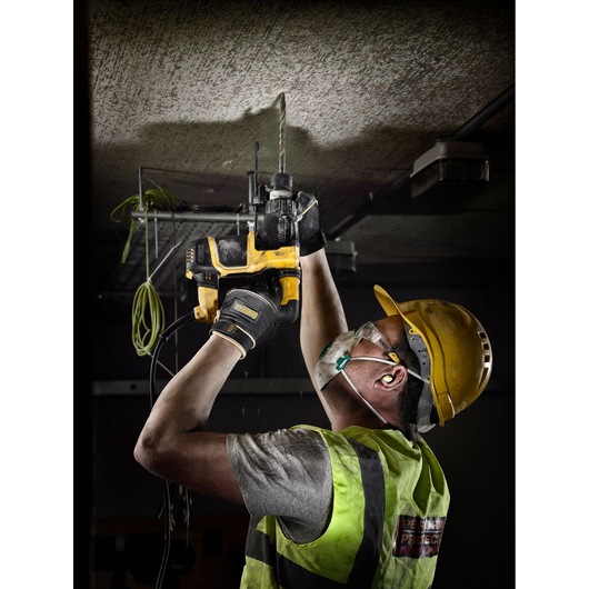 A construction worker wearing safety equipment, including a yellow hard hat, gloves, a protective vest, and a face mask, uses a DEWALT power drill to bore into a concrete ceiling.