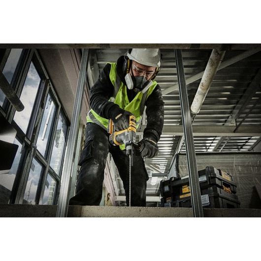 A construction worker wearing safety gear and a high-visibility vest is using a DEWALT power drill on a concrete surface at