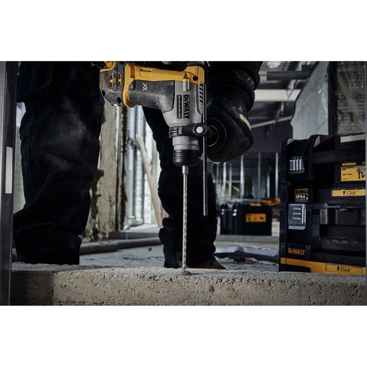 A DEWALT rotary hammer drill being used to drill into concrete at an indoor construction site. The tool is held by a person