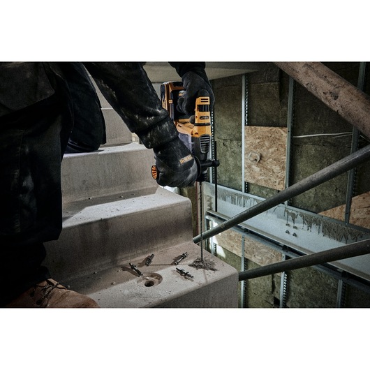 A worker wearing gloves is using a DEWALT power drill to drill into a concrete staircase in an unfinished building. Several