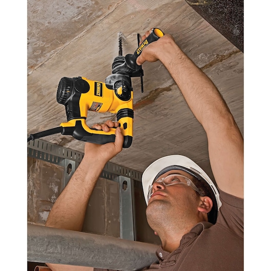 A person using a yellow and black DEWALT rotary hammer drill to drill into a concrete ceiling. The individual is wearing a brown shirt and a white safety helmet.