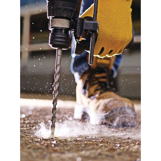 Close-up of a person wearing yellow work gloves and brown construction boots using a DEWALT drill with a masonry bit to drill into a concrete surface, with dust and debris visible.