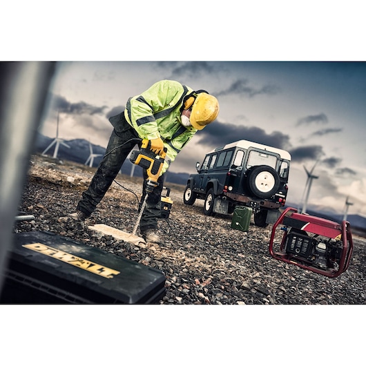 A construction worker wearing a yellow hard hat, green high-visibility jacket, and protective mask is using a DEWALT power drill to drill into the ground at an outdoor, rocky construction site. There is a black toolbox labeled DEWALT in the foreground, a red generator, and a black SUV with wind turbines and mountains in the background.