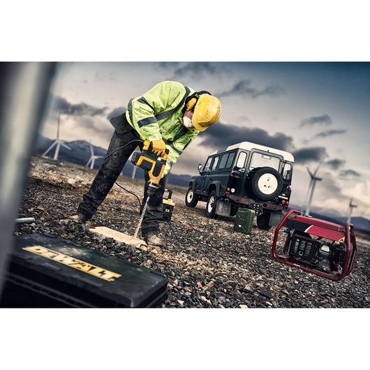 A construction worker wearing a yellow helmet and reflective jacket is using a DEWALT power drill on rocky ground outdoors. There is a toolbox with the DEWALT logo, a portable generator, a green fuel container, and an off-road vehicle in the background, with wind turbines and mountains in the distance under a cloudy sky.