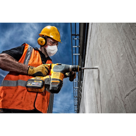 A construction worker wearing a yellow hard hat, orange safety vest, and gloves is using a DEWALT XR cordless rotary hammer