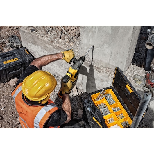 Construction worker wearing a yellow helmet and orange safety vest uses a DEWALT power drill to drill into a concrete wall
