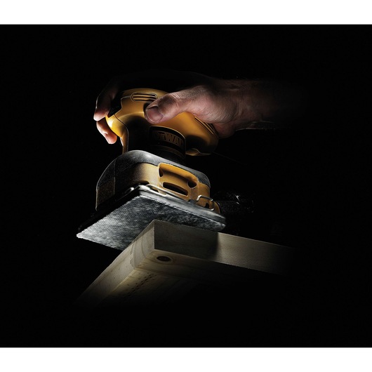 A close-up of a DEWALT hand sander being used on a piece of wood in a dark background.