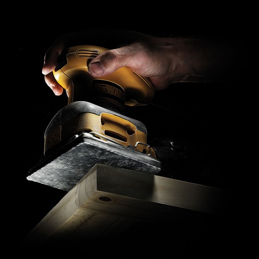 A close-up of a hand using a yellow and black DEWALT electric sander on a wooden plank against a dark background.