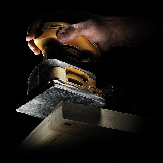A DEWALT electric sander held in a hand, sanding the edge of a wooden board against a dark background.