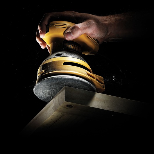 A close-up of a hand holding a yellow DEWALT orbital sander, smoothing the edge of a wooden plank against a black background.