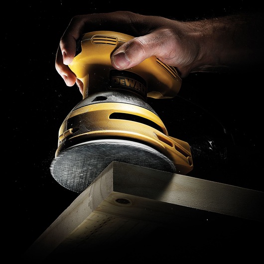 A close-up view of a hand holding a yellow DEWALT orbital sander, sanding the edge of a wooden plank.