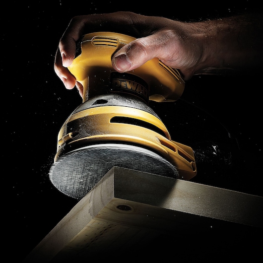 A close-up view of a hand holding a yellow DEWALT orbital sander, sanding the edge of a wooden board against a dark