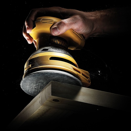 A close-up shot of a yellow DEWALT power sander being used on the edge of a wooden plank, with a hand gripping the tool