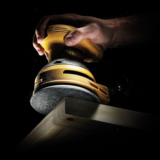 A close-up view of a hand using a DEWALT electric sander on the edge of a wooden board, with dust flying as the sander