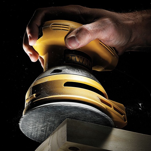 A close-up view of a person's hand using a DEWALT yellow orbital sander on the edge of a wooden block, with dust visible