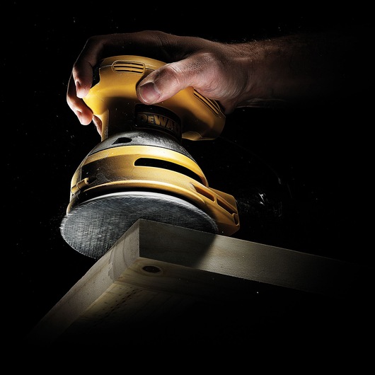 A close-up of a hand using a yellow DEWALT power sander on the edge of a wooden plank against a black background.