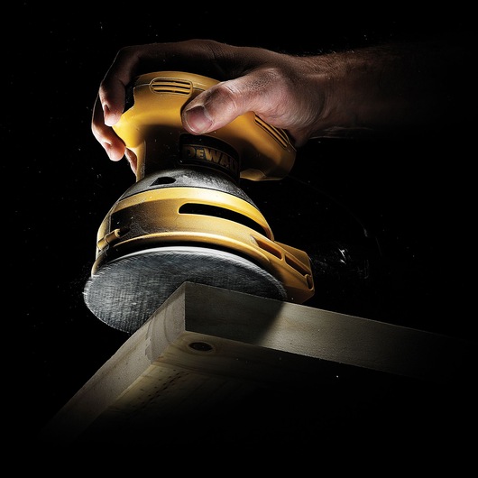 A hand holding a yellow DEWALT random orbital sander, smoothing the edge of a wooden plank against a dark background.