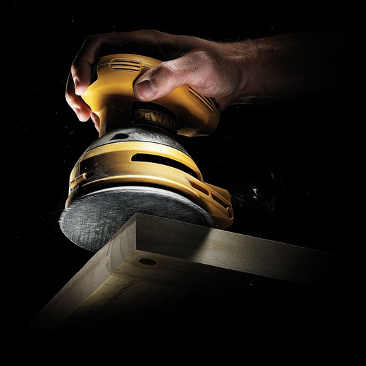 A close-up of a hand using a yellow DEWALT electric sander on a wooden surface, with the sander working on the edge of the