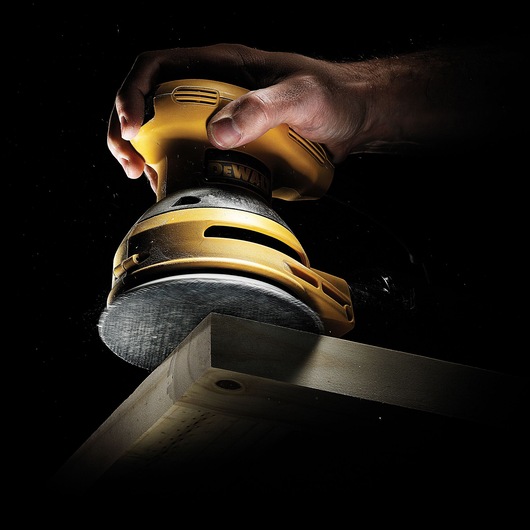 A close-up image of a hand operating a yellow DEWALT orbital sander on the edge of a wooden plank against a dark background.