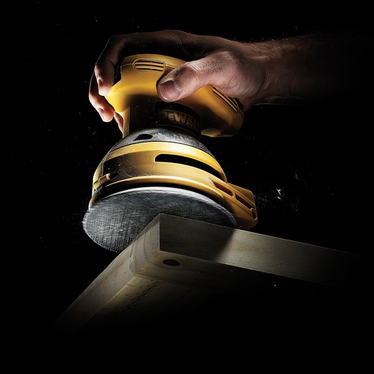 Hand using a yellow DEWALT sander to smooth the edge of a wooden board against a dark background.