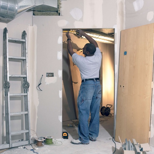 A person using a DEWALT DW0822CG laser level during an indoor construction or renovation project. The person is adjusting something near an air duct above a doorway. The scene includes a metal ladder, electrical wires, spools of cable, a wooden door, and construction materials scattered on the floor.