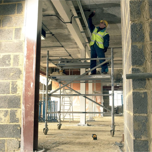 A construction worker wearing a safety vest and helmet stands on a mobile scaffold inside an unfinished building, reaching up to work on the ceiling. A yellow DEWALT laser level is positioned on the ground below.
