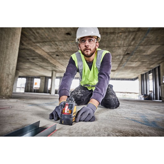 A person wearing a hard hat, safety vest, and gloves is kneeling on the floor of a construction site, using a DEWALT DW088K laser level. The device is projecting a visible red laser line for alignment. The surroundings show an unfinished building with concrete columns and open windows.