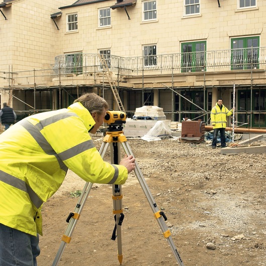 A person in a yellow high-visibility jacket is using a DEWALT spirit level on a tripod at a construction site, with another