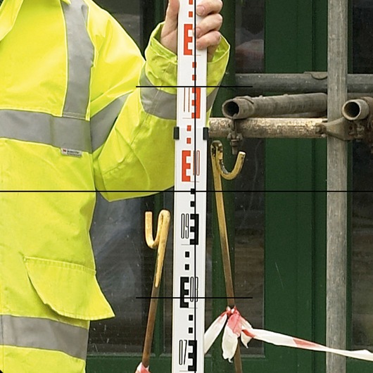 Person in a high-visibility jacket holding a survey staff next to construction tools and barriers.