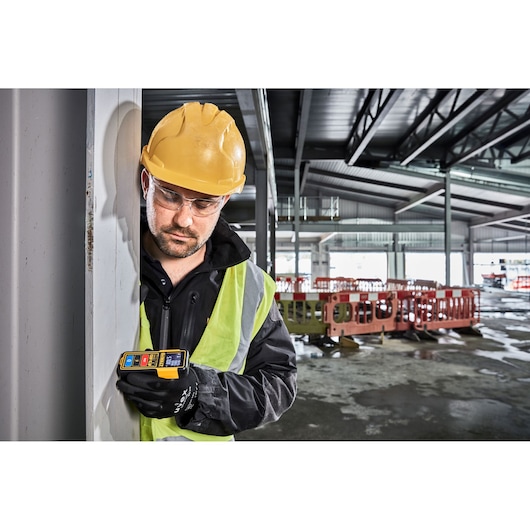 A construction worker wearing a yellow hard hat, high-visibility vest, and gloves is using a DEWALT DW099S laser distance