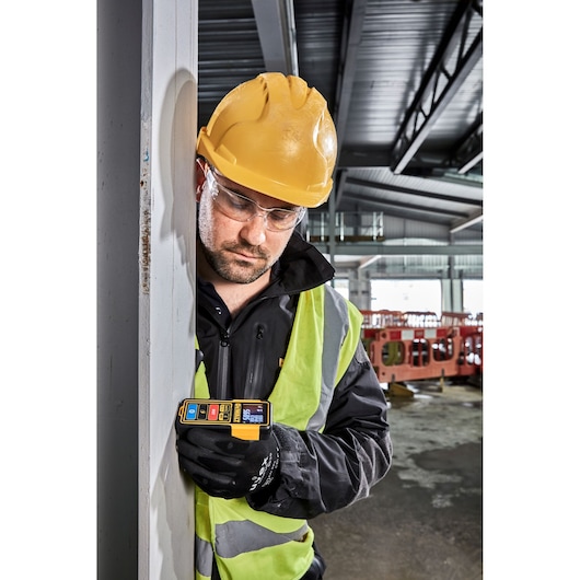 A construction worker wearing a yellow hard hat and reflective vest holds a DEWALT DW099S laser distance measurer against a