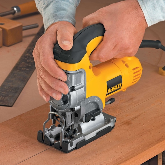 A close-up image of hands operating a yellow and black DEWALT jigsaw cutting through a piece of wood on a workbench, with woodworking tools visible in the background.