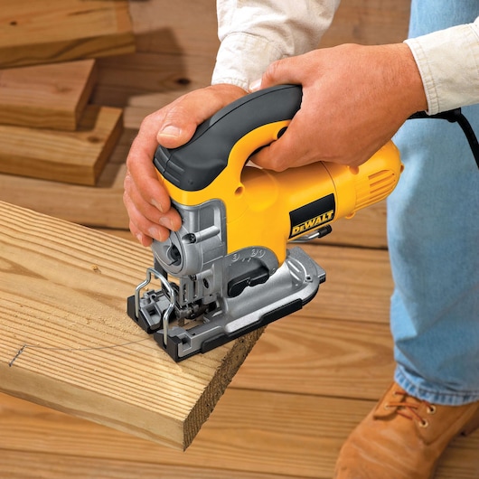 A person using a DEWALT DW331K jigsaw to cut a piece of wood. The jigsaw is yellow and black and is held firmly with both hands. The scene shows wooden boards and flooring.
