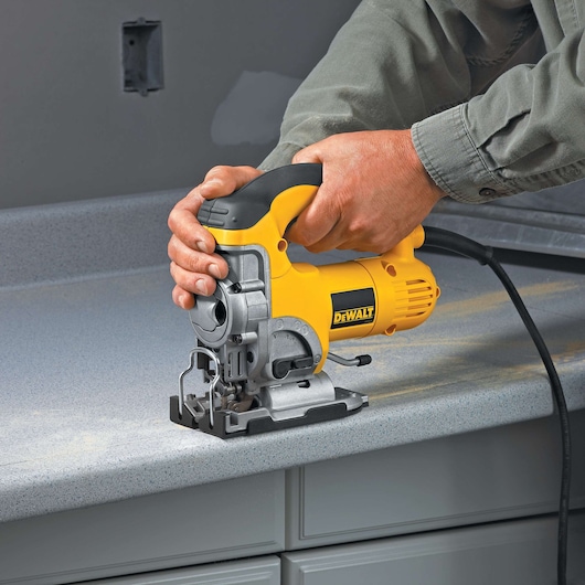 A person using a DEWALT electric jigsaw to cut a countertop. The yellow and black tool is being operated with both hands and is plugged into a power source.