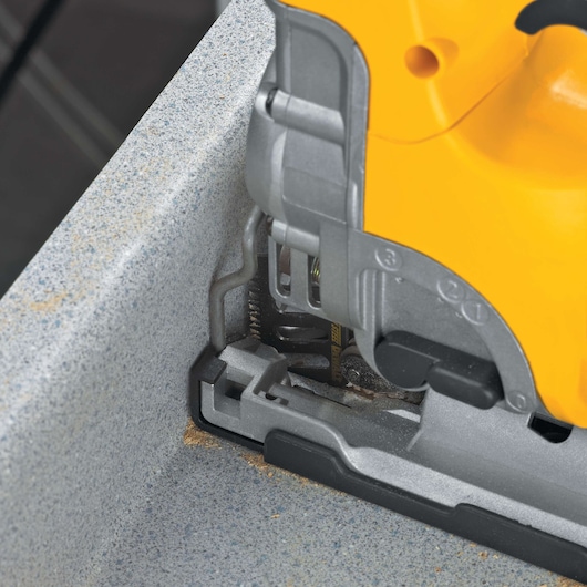 Close-up of a DEWALT jigsaw cutting through a gray textured surface, showing the tool's blade and base in action.