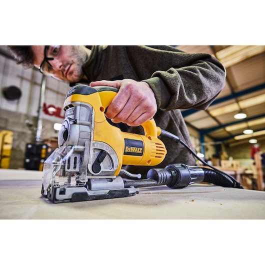A close-up view of a person using a DEWALT DW331K jigsaw to cut wood in a workshop. The yellow and black power tool is in action, with the person's hand firmly gripping it. The background shows an industrial workshop setting with wood and various tools.
