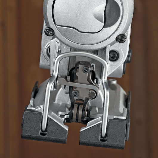 Close-up view of a metal jigsaw tool, focusing on the blade securing mechanism and protective guard, with a blurred wooden background.